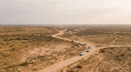 Vue aérienne symbolique d'une chaîne d'approvisionnement humanitaire traversant un paysage aride et fragmenté, évoquant la difficulté d'acheminer l'aide jusqu'aux populations les plus vulnérables.